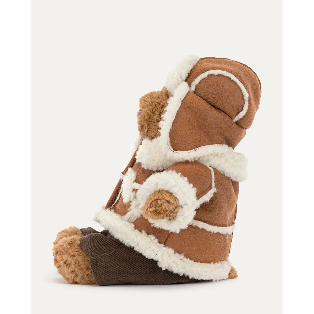 A plush teddy bear dressed in a brown and cream faux-shearling aviator-style jacket and matching hat, with dark brown corduroy pants, photographed on a clean off-white background.