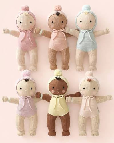 A knitted baby doll with a beige body and a light pink hooded outfit with a bow