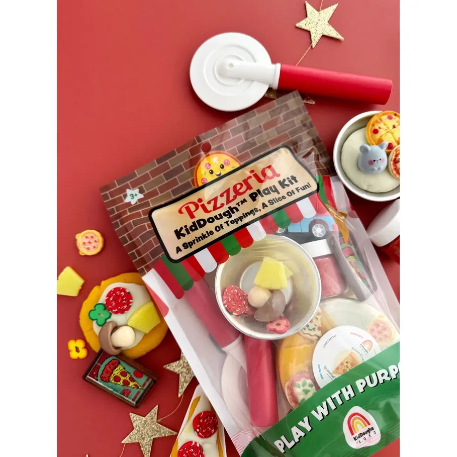 Pizzeria KidDough play kit in a clear plastic bag with a brown brick-pattern label, shown with toy pizza dough, red rolling pins, a small red and blue toy pizza truck, and assorted play food pieces on a white background.