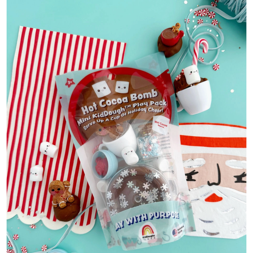 Hot Cocoa Bomb Mini KidDough Play Pack shown in its retail bag with the contents spilled out in front, including a clear cup, marshmallows, a gingerbread figure, candy canes, sprinkles, and a hot cocoa cup on a white background.