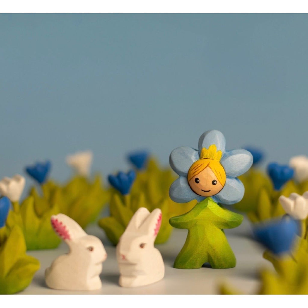 Toy figure with a blue flower head and green body on a wooden surface with a light blue background