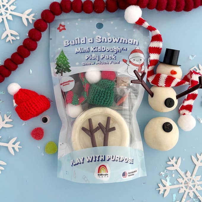 Build a Snowman Mini McDough Play Pack toy set in a clear plastic bag, with assembled snowman figures, a red knit hat, and other winter-themed play pieces displayed in front on a white background.