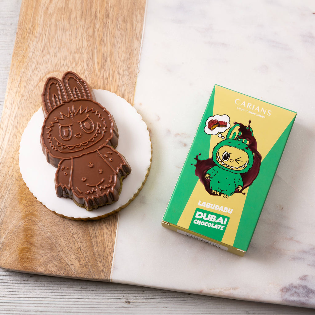 Chocolate rabbit-shaped cookie on a white plate with a green and yellow box labeled 'Dubai Chocolate' featuring a cartoon character.
