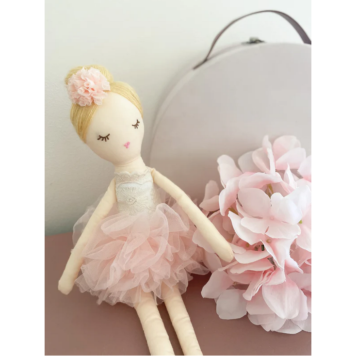 Handmade doll in a pink dress with a flower in its hair next to pink flowers on a neutral background