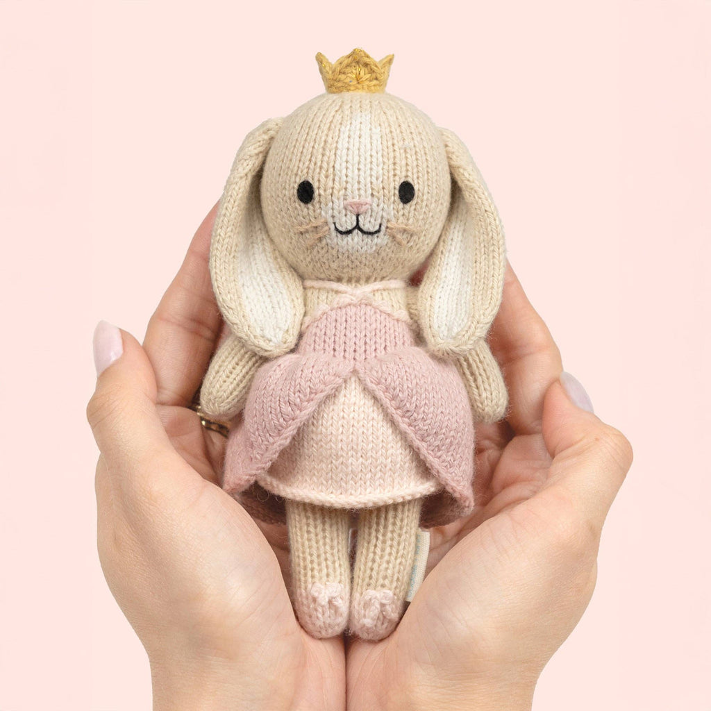 Knitted bunny toy with a pink dress and crown in front of a floral-patterned box on a soft pink background.