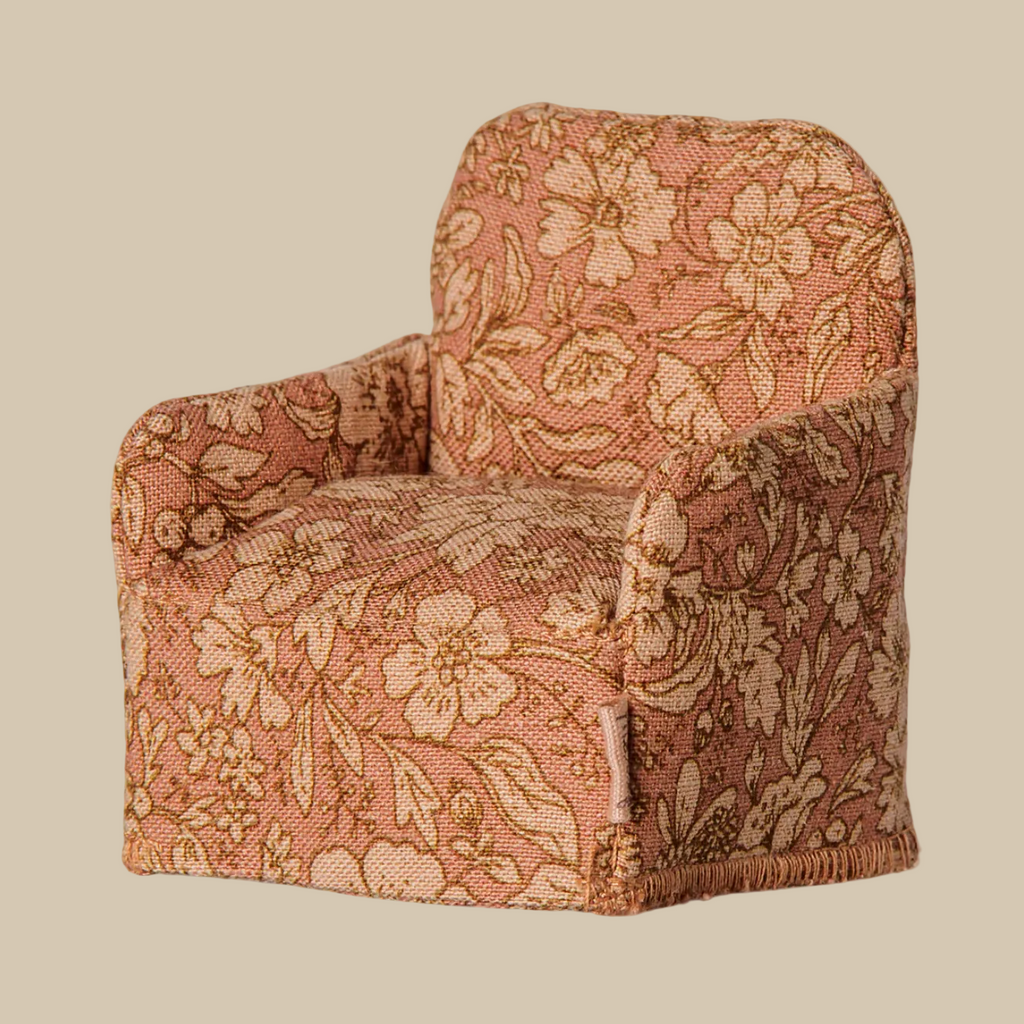 A small upholstered armchair with a warm floral pattern in dusty pink, beige, and tan, shown against a plain light beige background. A small fabric tag on the side reads Maileg