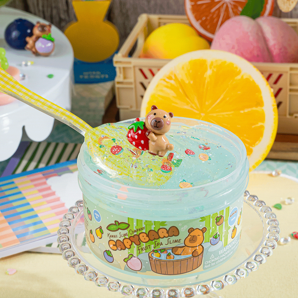 A round plastic container of Kawaii Slime Company Capybara Fruit Spa Slime, featuring a clear domed lid with three small bear figurines and colorful confetti, tied with a large green ribbon bow. The container label shows a cartoon Capybara in a bath with fruit illustrations.