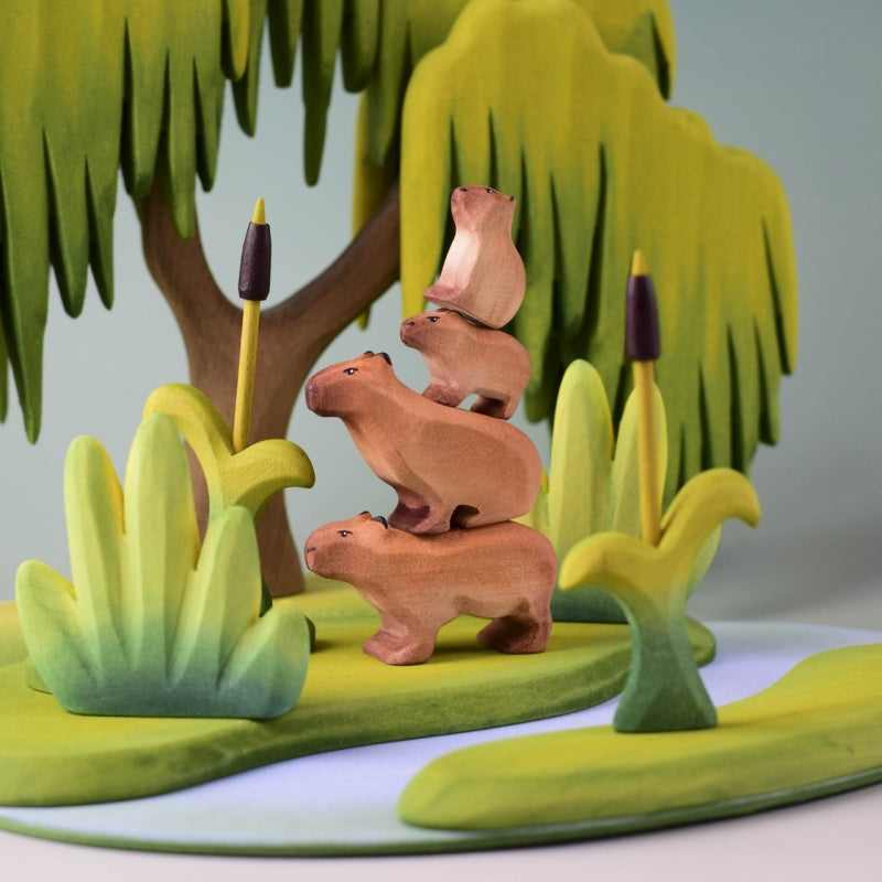 Four small wooden capybara figurines in natural light brown wood tones, arranged on a light wood surface against a soft light blue background.