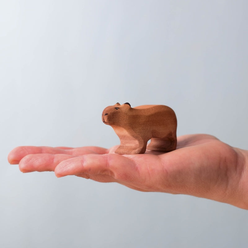 A small hand-carved wooden animal figurine, shaped like a capybara, standing on a light wood surface with a soft blue background.