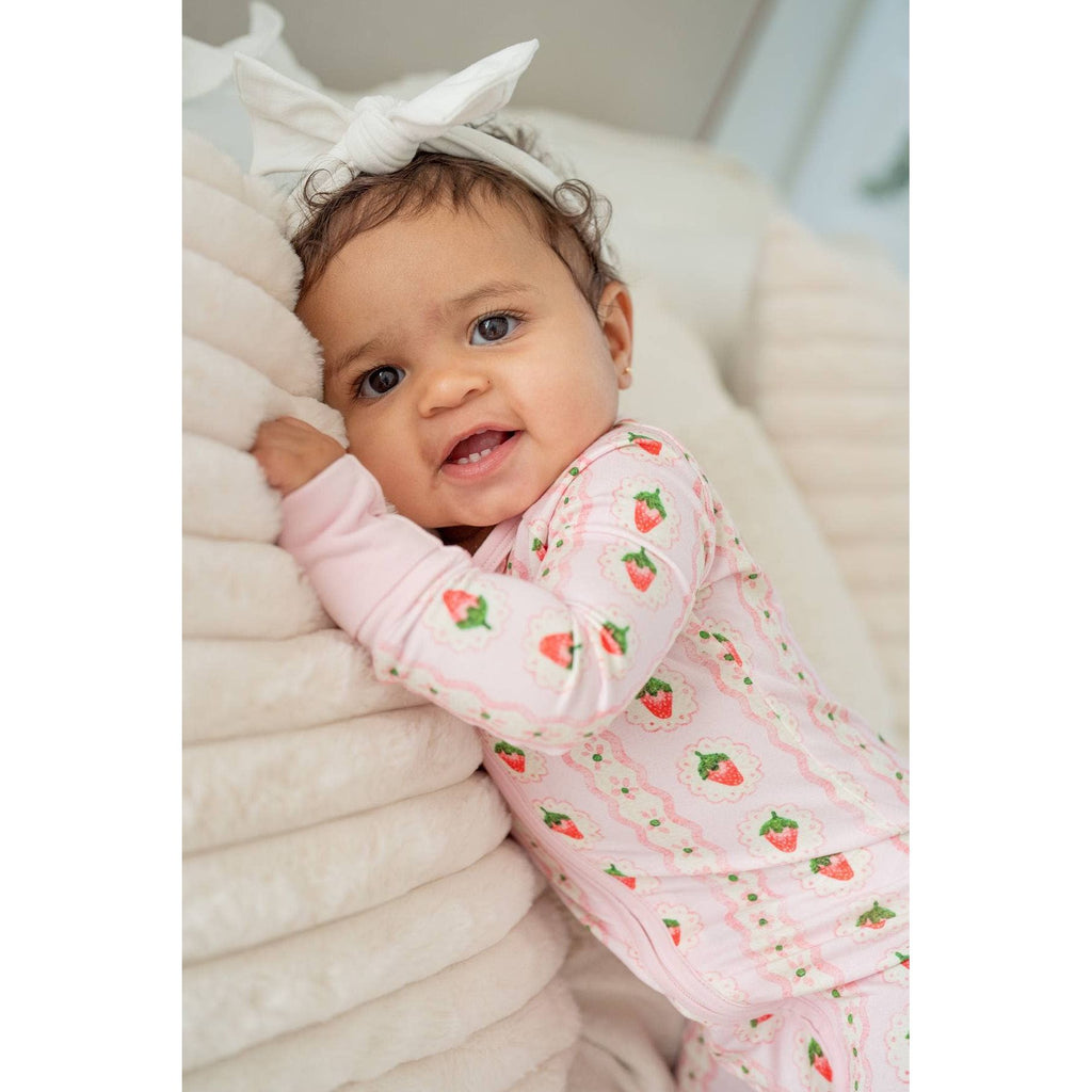 Baby wearing a pink strawberry print outfit with a white headband, sitting on a couch.