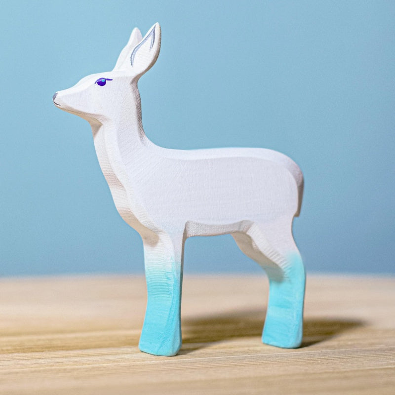 A stylized wooden deer figurine with a white body and light blue gradient legs, standing on a wooden surface against a soft blue background.