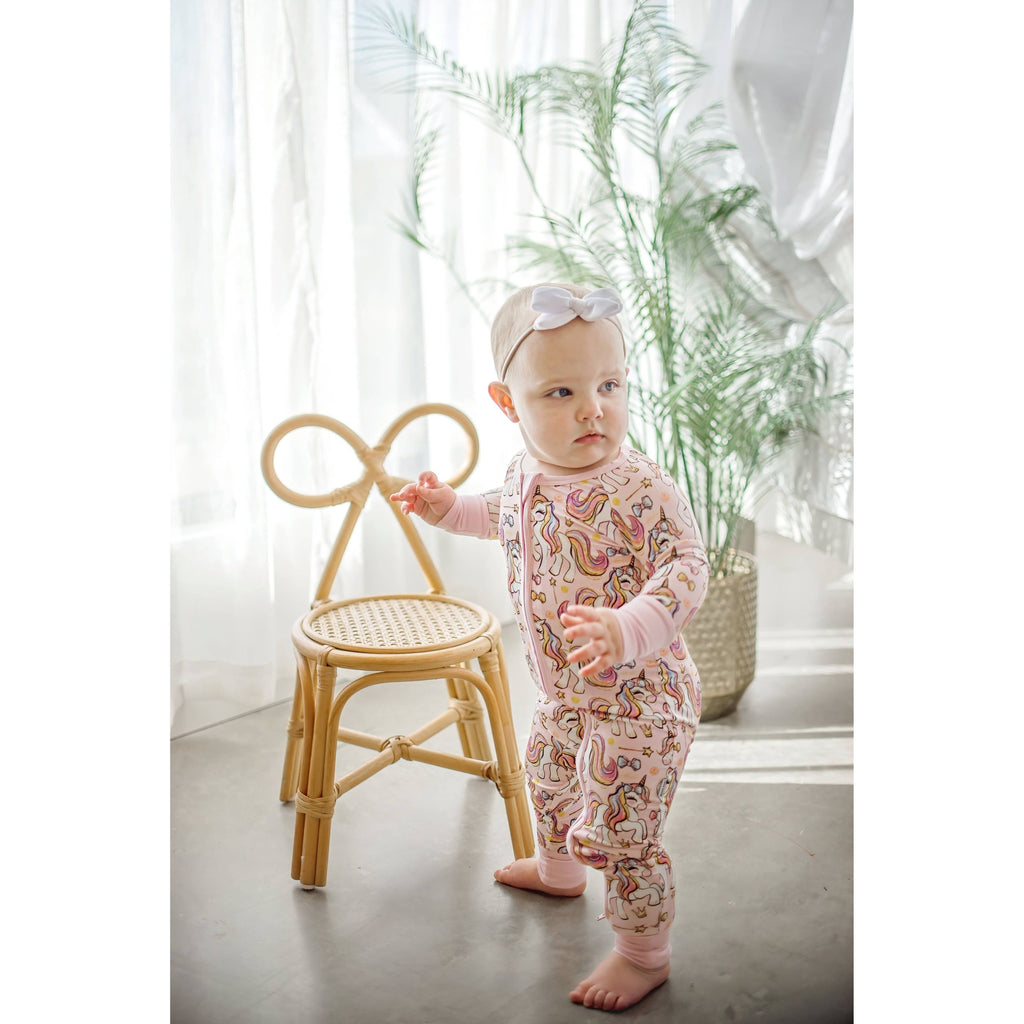 Baby in a pink unicorn onesie sitting on a small stool with white curtains in the background.