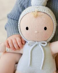 A knitted baby doll with a pale peach face and limbs, wearing a light blue hooded romper with a bow