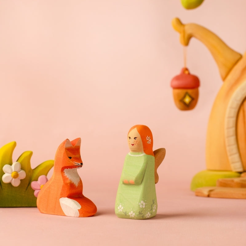 A small hand-painted wooden angel figurine with orange hair and a light green dress, standing on a light wooden surface against a soft mint green background.