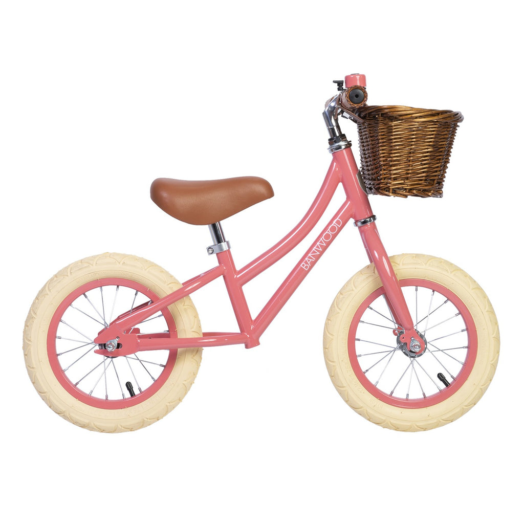 A pink balance bike with a brown saddle, matching grips, and a front wicker basket. The bike is designed for young children.