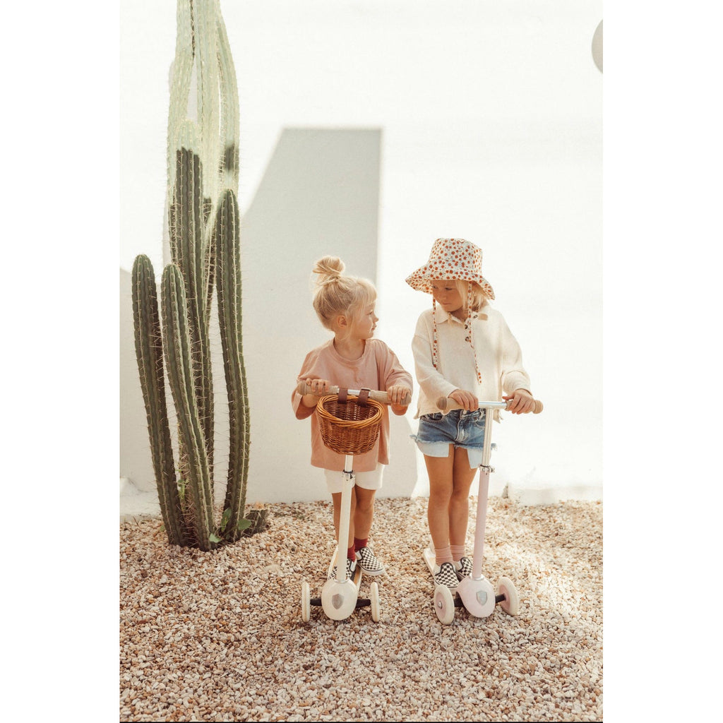Two children standing side by side on a Banwood three-wheeled scooter with a cream-colored frame. The scooter has a wide deck with black rubber grip tape, and two front wheels and one rear wheel.