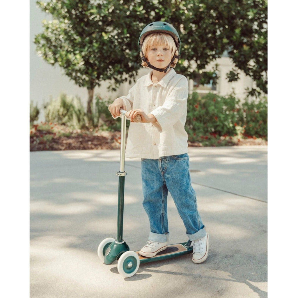 A child riding a green three-wheeled scooter with a wide deck and a T-handlebar, outdoors.