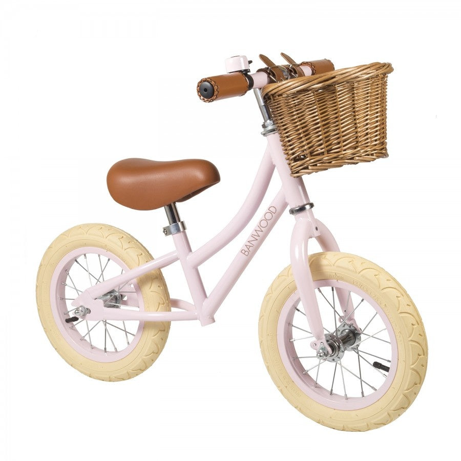 A pink balance bike with a brown saddle, white frame, and a wicker basket in front.