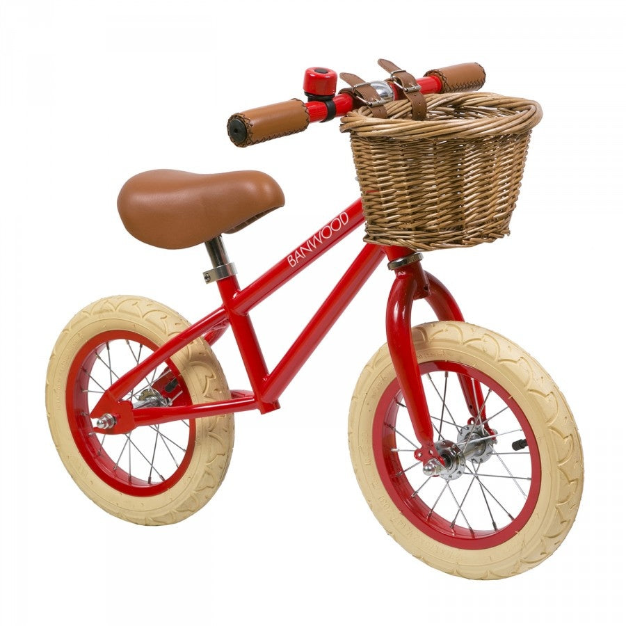 A red balance bike with a white frame, brown saddle, and a wicker basket on the front. The bike has large wheels and is designed for young children.
